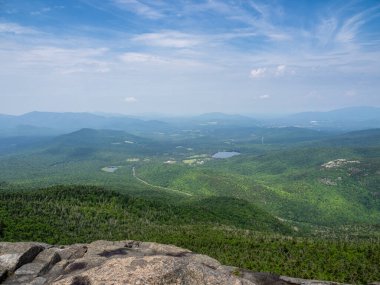 Cascade Dağı 'nın zirvesinden nefes kesici bir manzara, engebeli bir kaya ön planı ve New York' taki Lake Placid 'den görülebilen Olimpiyat Kayak Kompleksi.