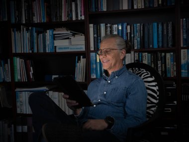 With a warm glow on his face, a man in a blue shirt smiles while reading on a tablet, surrounded by books on a cozy bookshelf. clipart