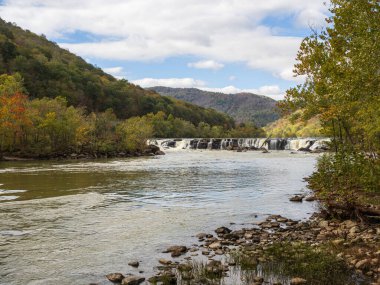 Sandstone Falls, New River Gorge Ulusal Parkı, Batı Virginia, parlak mavi bir gökyüzü altında parlak sonbahar renkleri arasında akar, dağları aydınlatan güneş ışınları ve ön planda dağılmış taşlar, doğanın canlı güzelliğini yakalar..