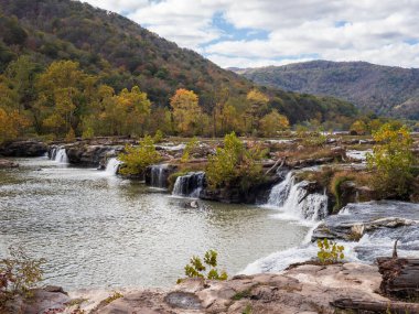 Batı Virginia 'daki New River Gorge Ulusal Parkı' ndaki Sandstone Şelaleleri, arka planda rengarenk dağ yamaçlarıyla güçlü bir şekilde akar ve doğanın canlı mevsimsel güzelliğini gösterir..