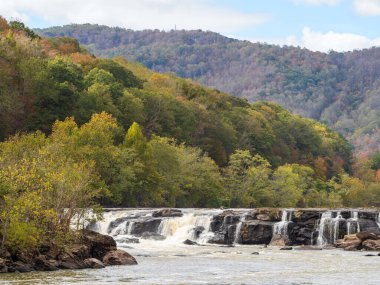 Sandstone Şelalesi, New River Vadisi Ulusal Parkı, Batı Virginia, parlak sonbahar renklerinde parlak mavi bir gökyüzü altında çağlarken, güneş ışınları dağ yamaçlarını sıcak, altın bir ışık altında yıkıyor..