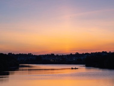 Canlı bir günbatımı, iki kişilik küçük bir balıkçı teknesinin yer aldığı dingin bir göl üzerinde ılık renkler oluşturur..