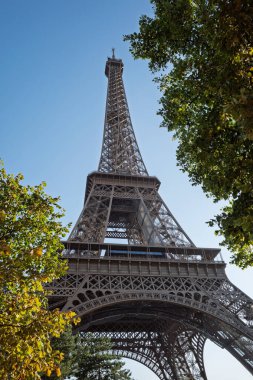 Muhteşem Eyfel Kulesi açık mavi gökyüzüne karşı canlı yeşil ağaçlar tarafından çerçevelenmiş ve Paris, Fransa 'nın eşsiz bir perspektifinden ihtişamını yakalamıştır..