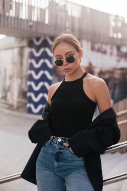Moda kıyafetleri olan bir model. Şehir manzarası. Gençlik modelliği. Şehir modası trend sahnesi. Sokak modası. Siyah ceketli, kot pantolonlu ve güneş gözlüklü muhteşem bir kadın. 