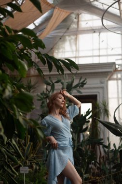 Mavi elbiseli genç kadın, arka planda yeşillik olan tropik botanik bahçesinde poz veriyor.