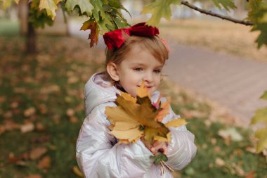 Kafasında kırmızı bir fiyonk ve elinde sonbahar yaprakları olan küçük bir kız..