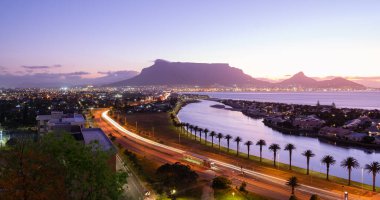 Masa Dağı 'nın akşam manzarası ve Cape Town şehrinin aydınlanması, günbatımı renklerini yansıtan lagün ve ön planda Woodbridge Adası, Milnerton, Güney Afrika