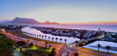 Woodbridge Adası ve alacakaranlıkta birleşen köprüsü, deniz feneri dimdik dururken ve gölcük günbatımının sıcak renklerini yansıtırken Milnerton, Cape Town, Güney Afrika