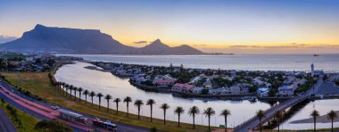 Woodbridge Adası ve alacakaranlıkta birleşen köprüsü, güneş Masa Dağı ile Milnerton, Cape Town 'daki Lion' s Head Dağı arasında batarken deniz feneri ve gölü yansıtan gölle birlikte. Güney Afrika