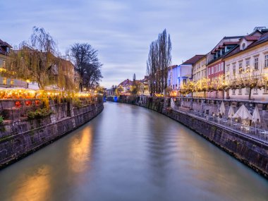 Slovenya 'nın Ljubljana Nehri boyunca alacakaranlıkta aydınlanan Noel ışıkları ve eski kasaba binaları