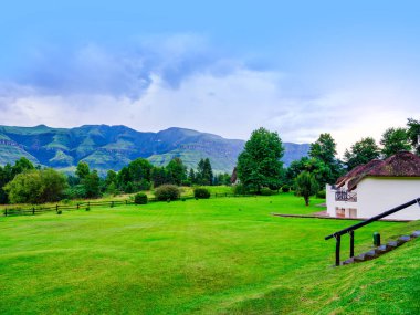 A thatch-roof house on the lush grass field with Drakensberg mountain in the background, KwaZulu-Natal, South Africa clipart