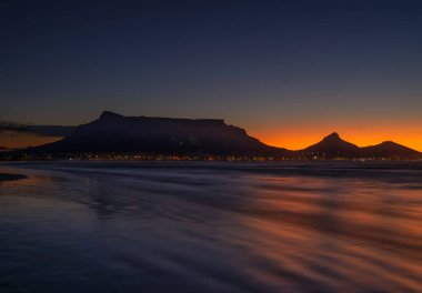 Gün batımından sonra Güney Afrika 'daki Sunset Beach' ten Masa Dağı silueti ve aydınlatılmış Cape Town 'un uzun pozlu görüntüsü.