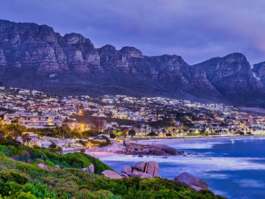 Günbatımından sonra Kamp Koyu 'nun canlı ışıklarını, 12 Havarinin görkemli dağ sırasını dramatik bir zemin olarak yansıtan güzel bir uzun pozlama. Cape Town, Güney Afrika