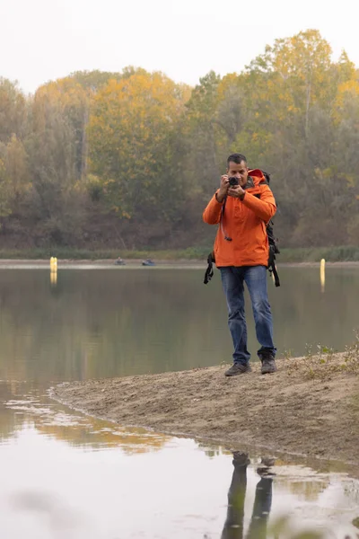Portrait of traveler man at lake in autumn taking a photo with the digital camera and orange jacket. High quality photo