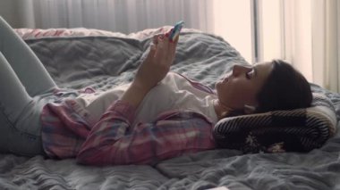 Evdeki kanepede uzanmış akıllı telefon kullanan beyaz bir kadın. Evdeki bir yatakta kablosuz kulaklıkla müzik dinleyen bir kız.