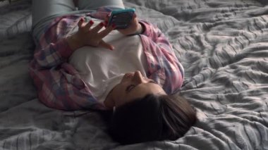 Evdeki kanepede uzanmış akıllı telefon kullanan beyaz bir kadın. Evdeki bir yatakta kablosuz kulaklıkla müzik dinleyen bir kız.