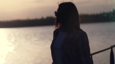 Uzun saçlı ve güneş gözlüklü zarif bir kadının silueti rüzgarlı bir günde gün batımında kıyıda duruyor ve suya bakıyor..