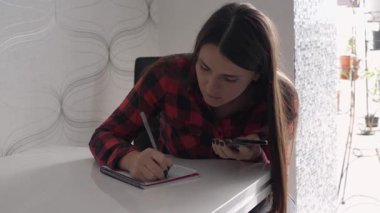 Focused young woman sitting at desk, writing notes in a notebook while checking smartphone. Concept of remote work, planning, productivity, studying or self-organization. 