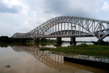 İkonik jübile köprüsü Hooghly Nehri boyunca uzanır, klasik çelik kemer bağı tasarımı, görüntü köprünün çarpıcı geometrik simetrisini yakalar ve ön planda yeşillik ve dramatik bulutlu gökyüzü ile muson köpüklü nehrin yansıması