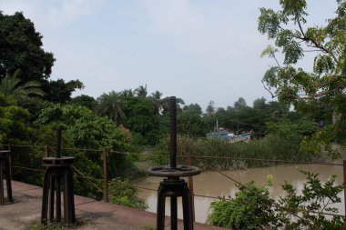 Geleneksel su kontrol mekanizmaları Geokhali 'deki nehir kıyısında vida mekanizmaları, yoğun yeşillik, palmiye ağaçları ve uzakta bir balıkçı teknesi, kırsal geçim, altyapı ve doğal manzara karışımı sakin bir nehir manzarasına bakıyor