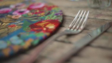 Bıçak ve çatal, ahşap masada, renkli bir el yapımı paspasın yanında. Yavaş çekim, yüzeysel alan derinliği. 