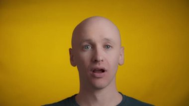 Mavi gözlü ve parlak tenli bir adam kameraya doğru bakıyor ve sarı arka planda esniyor..