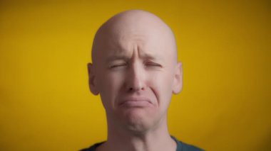Mavi gözlü ve parlak tenli bir adam kameraya doğru bakıyor ve sarı arka planda ağlayan bir yüz yapıyor..