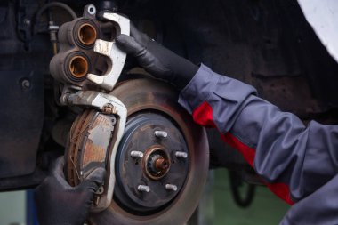 Bir araba servisindeki tamirci arabanın ön tarafındaki kalibre ve fren pedlerini değiştiriyor. Tekerleksiz araba. Metal yedek parçalar. Fren diskini iki eliyle tutuyor.