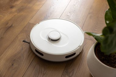 Süpürge fırçası, hareket sensörleri ve kamera ile beyaz bir robot süpürge, çiçek saksısının yakınındaki ahşap zemini temizler ve odaya dokunmaz. aygıt gövdesindeki kontrol düğmeleri.