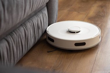 Beyaz bir robot süpürge yerdeki koyu meşe ağacını temizliyor. Zor yerlerde kanepenin yanında toz topluyor. Modern teknolojilerin otomatik yönetimi. Kızılötesi sensör kontrolü 