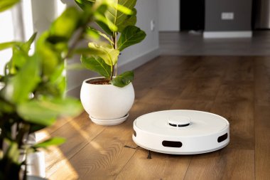 Süpürge fırçası, hareket sensörleri ve kamera ile beyaz bir robot süpürge, çiçek saksısının yakınındaki ahşap zemini temizler ve odaya dokunmaz. aygıt gövdesindeki kontrol düğmeleri.
