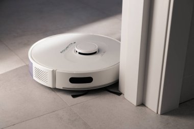 Beyaz bir robot elektrikli süpürge beyaz kapının yanındaki gri fayansları temizler. Toz toplar ve yerleri siler. Samimi bir dairede sabah. Odadaki güneş ışınları