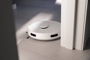 Beyaz bir robot elektrikli süpürge beyaz kapının yanındaki gri fayansları temizler. Toz toplar ve yerleri siler. Samimi bir dairede sabah. Odadaki güneş ışınları