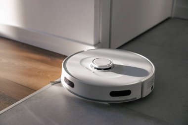 Beyaz bir robot süpürge engelsiz bir şekilde eşikten fayansları ve ahşap zeminleri temizliyor. Toz toplar ve yerleri siler. Samimi bir dairede sabah. Odadaki güneş ışınları