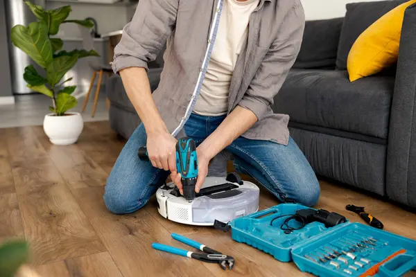 man repairs robot vacuum cleaner with tools and screwdriver sitting on floor wearing blue jeans and gray shirt. Scheduled service and cleaning in a room with a gray sofa and green flower pots