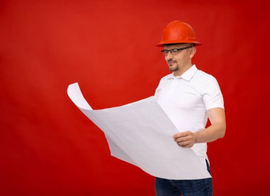 Beyaz tişörtlü ve kırmızı kasklı bir erkek mühendis inşaat planlı büyük beyaz bir çarşaf tutuyor. Kırmızı bir duvarın arkasında. Gözlüklü bir adam kameraya bakıyor.