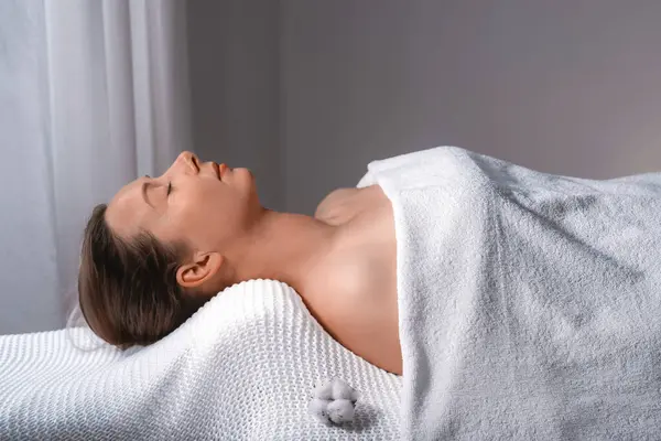 A woman lays on a massage table, covered in a white towel. Her body is relaxed, with her nose, leg, eyelash, jaw, ear, sleeve, gesture, elbow, and thigh visible