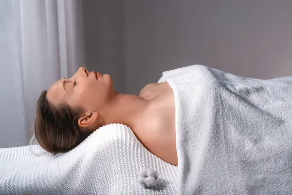 A woman lays on a massage table, covered in a white towel. Her body is relaxed, with her nose, leg, eyelash, jaw, ear, sleeve, gesture, elbow, and thigh visible