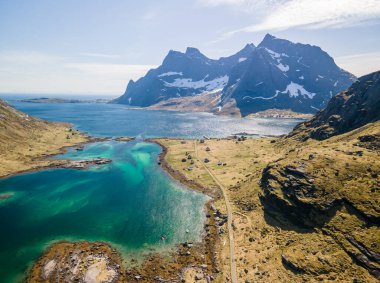 Lofoten, Norveç 'teki Vindstad köyünün insansız hava aracı görüntüsü, küçük renkli evler, büyük dağlar ve açık bir bahar gününde mavi bir göl.