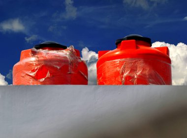 Bir binanın beton çatısındaki iki turuncu plastik su deposu. Arka plandaki beyaz bulutlar güneşli bir günde mavi gökyüzüne karşı.