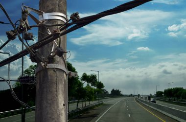 Kablolardan oluşan kaotik halat ve kablolarla dolu bir elektrik direği açık gökyüzünün olduğu bir günde paralı bir yolun arka planına karşı.