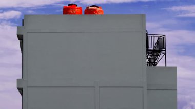 Arka planda mavi ve bulut hareketi olan iki su deposu tankı ve siyah demir merdivenli gri çok katlı bir bina.