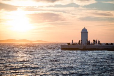 ZADAR, CROATIA - 11 AUG 2024: Deniz fenerinin çevresindeki insanlar Hırvatistan 'ın Zadar kentinde altın saat günbatımına hayran