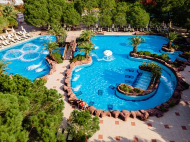 Lüks bir sahil otelinde geniş havuzlu ve slaytlı bir su parkı manzarası. Eğlenceli ve rahatlatıcı bir tatil için mükemmel..