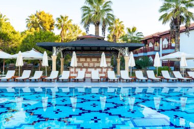 Şezlonglar ve şemsiyelerle dolu lüks bir tatil köyünde kristal berraklığında yemyeşil bir havuzun yanında mükemmel bir yaz günü bekliyor.
