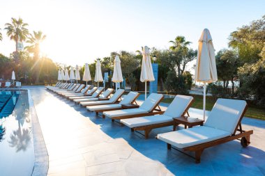 Boş şezlonglar ve şemsiyelerle dolu lüks bir tatil köyünde bereketli ağaçlar ve açık mavi gökyüzü ile çevrili el değmemiş bir yüzme havuzu.