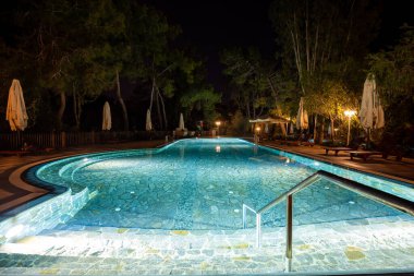 Boş güverte sandalyeleri ve düzgünce katlanmış şemsiyeleri olan akşam vahası güzel aydınlatılmış bir yüzme havuzu olan bir turizm beldesi.