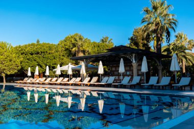 Şezlonglar ve şemsiyelerle dolu lüks bir tatil köyünde kristal berraklığında yemyeşil bir havuzun yanında mükemmel bir yaz günü bekliyor.