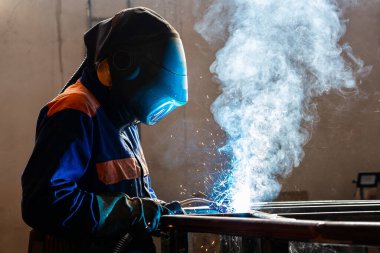 Fabrikada kaynak makinesiyle çalışan yetenekli bir metal işçisi güvenlik ekipmanı takıyor. El becerisi iş gücü kavramına göre metal imalatı ve inşaat bakım hizmeti