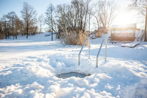 Idyllic winter swimming spot with hole and metal ladder on a snowy lakeside at sunrise